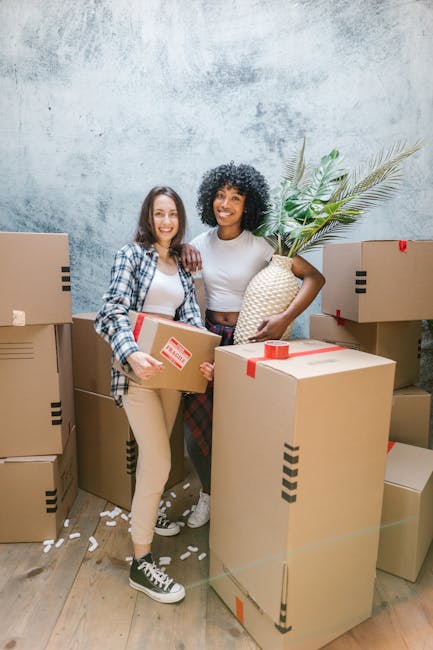 Close-up image of a person wearing a dark green shirt and blue trousers, carefully handling a large cardboard box during a home relocation process. The individual is gripping the box with both hands,